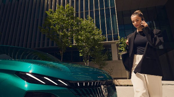 Woman talking on phone next to a teal car parked in front of a building with trees nearby.