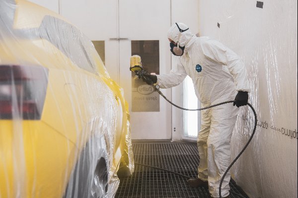 Technician in protective gear spray painting a covered yellow car.