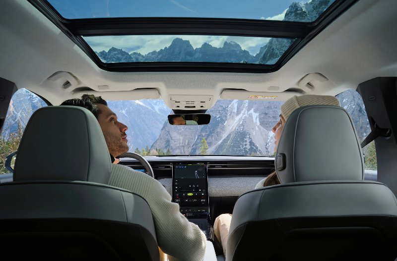 Two people in a car with panoramic sunroof facing a mountainous landscape.