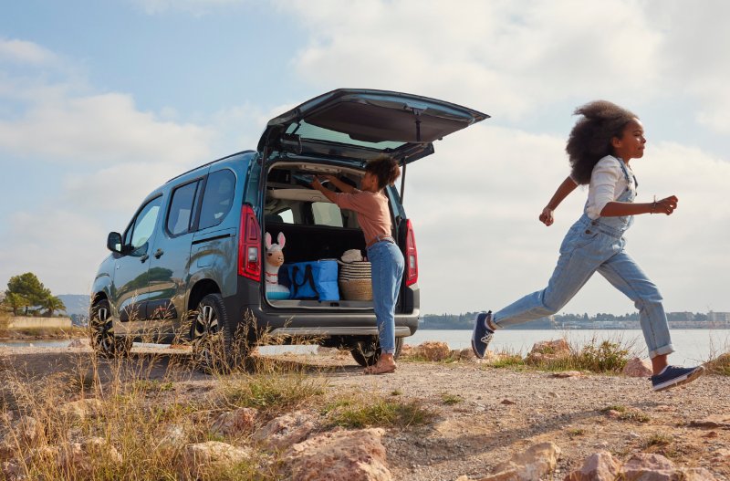 Child runs by an open car boot near a lake, with another person arranging items inside.
