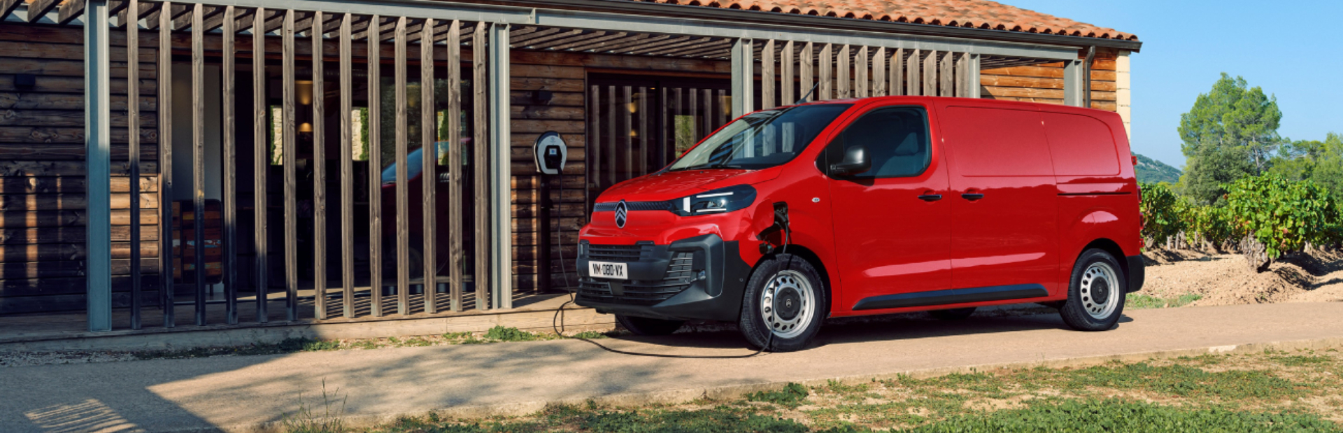 Red electric van charging outside a wooden building.