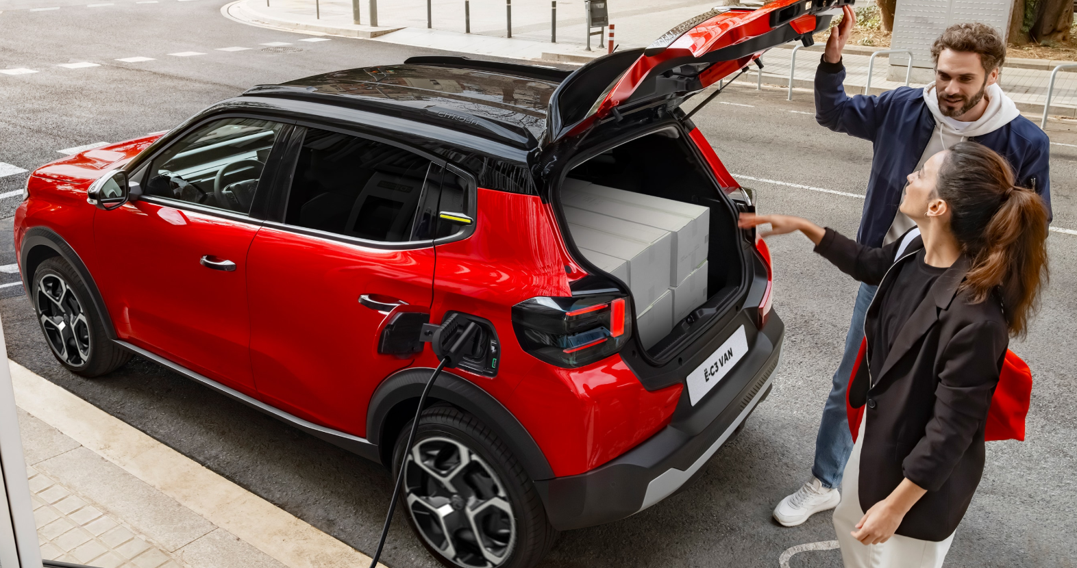 Red electric car charging with boot open, two people loading boxes.