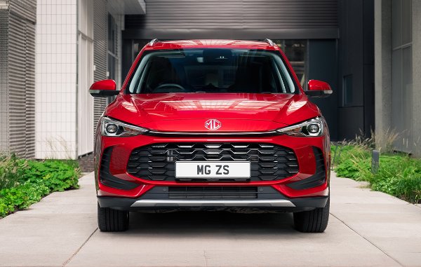 Red MG ZS car parked between buildings and greenery, viewed from the front.