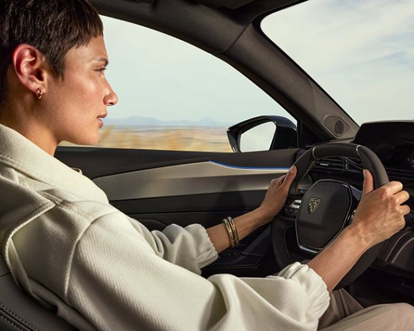Person driving a Peugeot car, viewed from the side, holding the steering wheel with both hands.