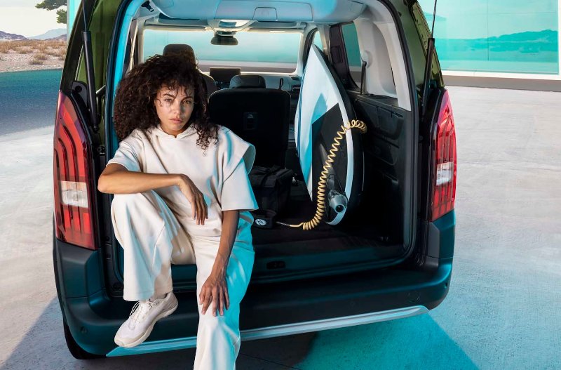 Person sitting in the back of a car with a surfboard next to them.
