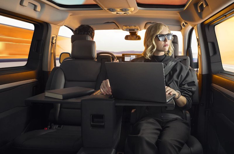 Person with a laptop and book in a car's back seat, with another person driving.