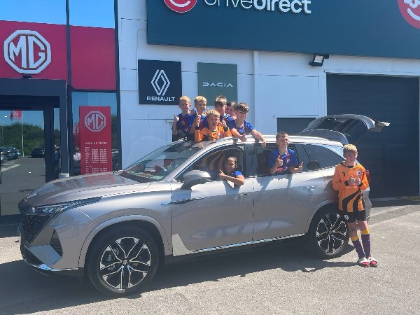 Children in football kits posing in and around a silver MG car outside a dealership.