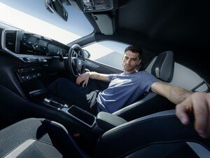 Man sitting inside a car, reaching towards the camera, with the steering wheel visible.