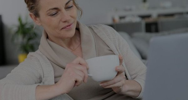 Woman in a beige sweater holding a coffee cup and looking at a laptop screen.
