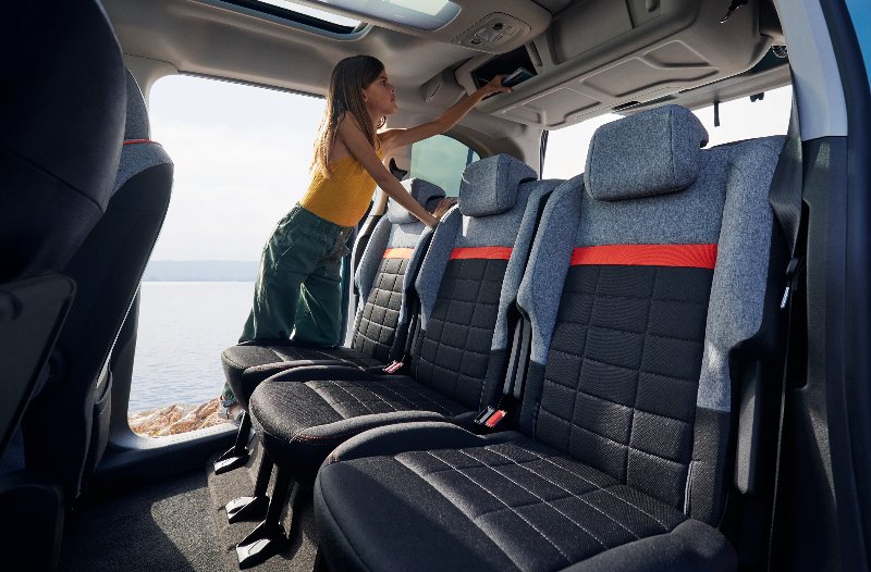 Woman standing in a car adjusting something above the rear seats with grey and red upholstery.
